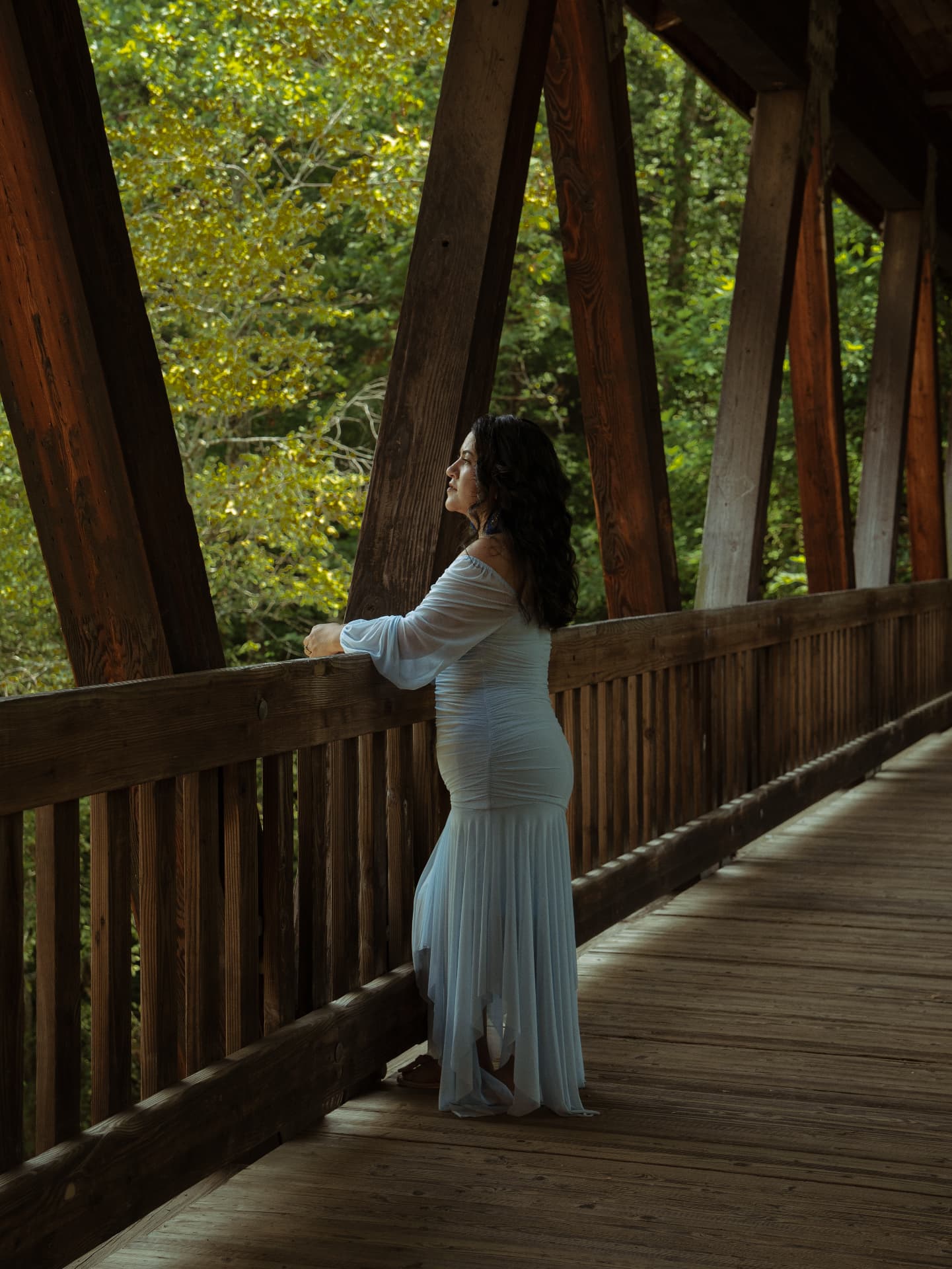 Portrait of a woman in a blue gown looking out from a covered bridge into the trees