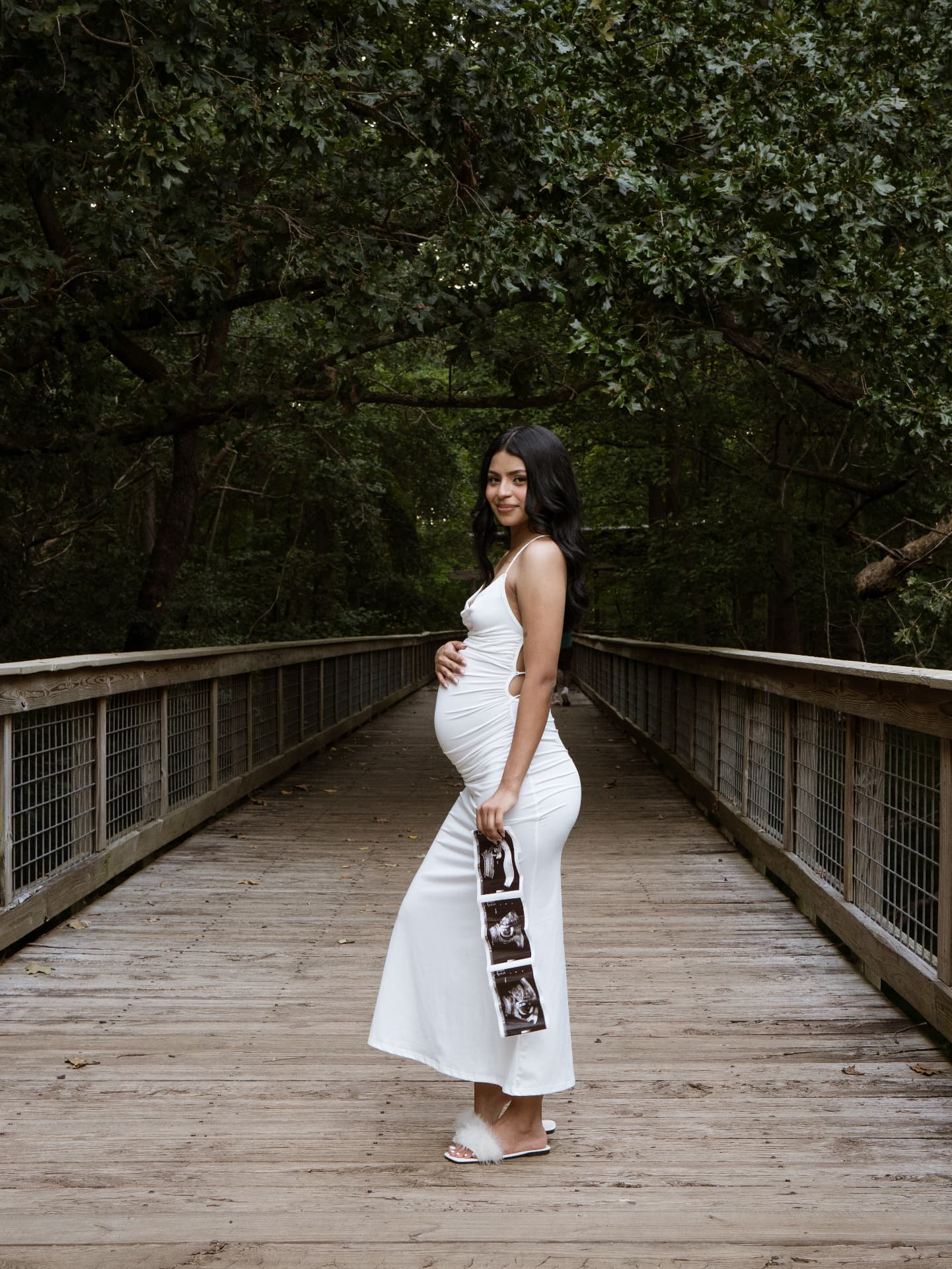 Expecting mother in a white gown holding ultrasound photos on a wooded footbridge