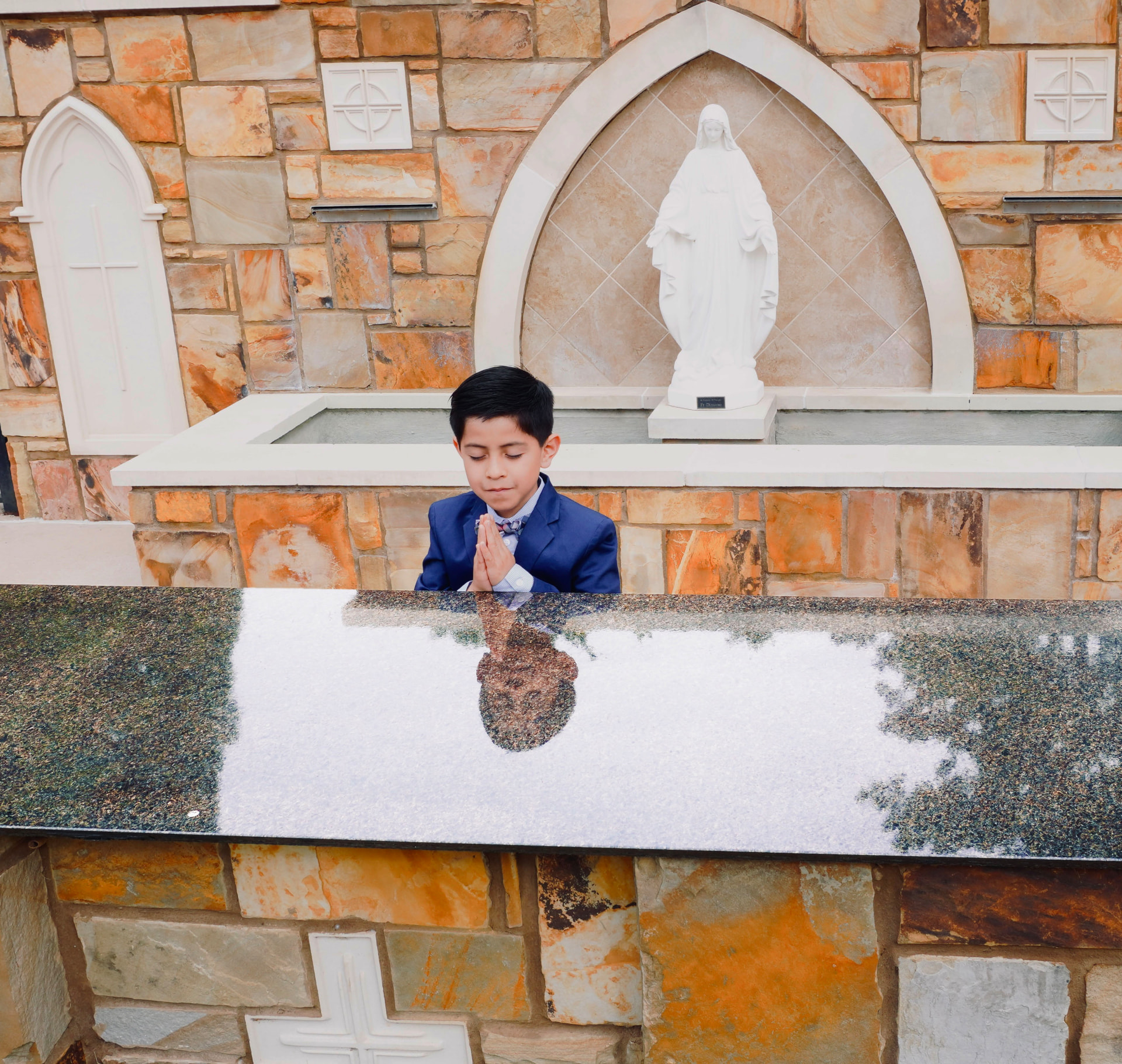 Boy in a blue suit praying beside a stone chapel and Marian statue in Georgia