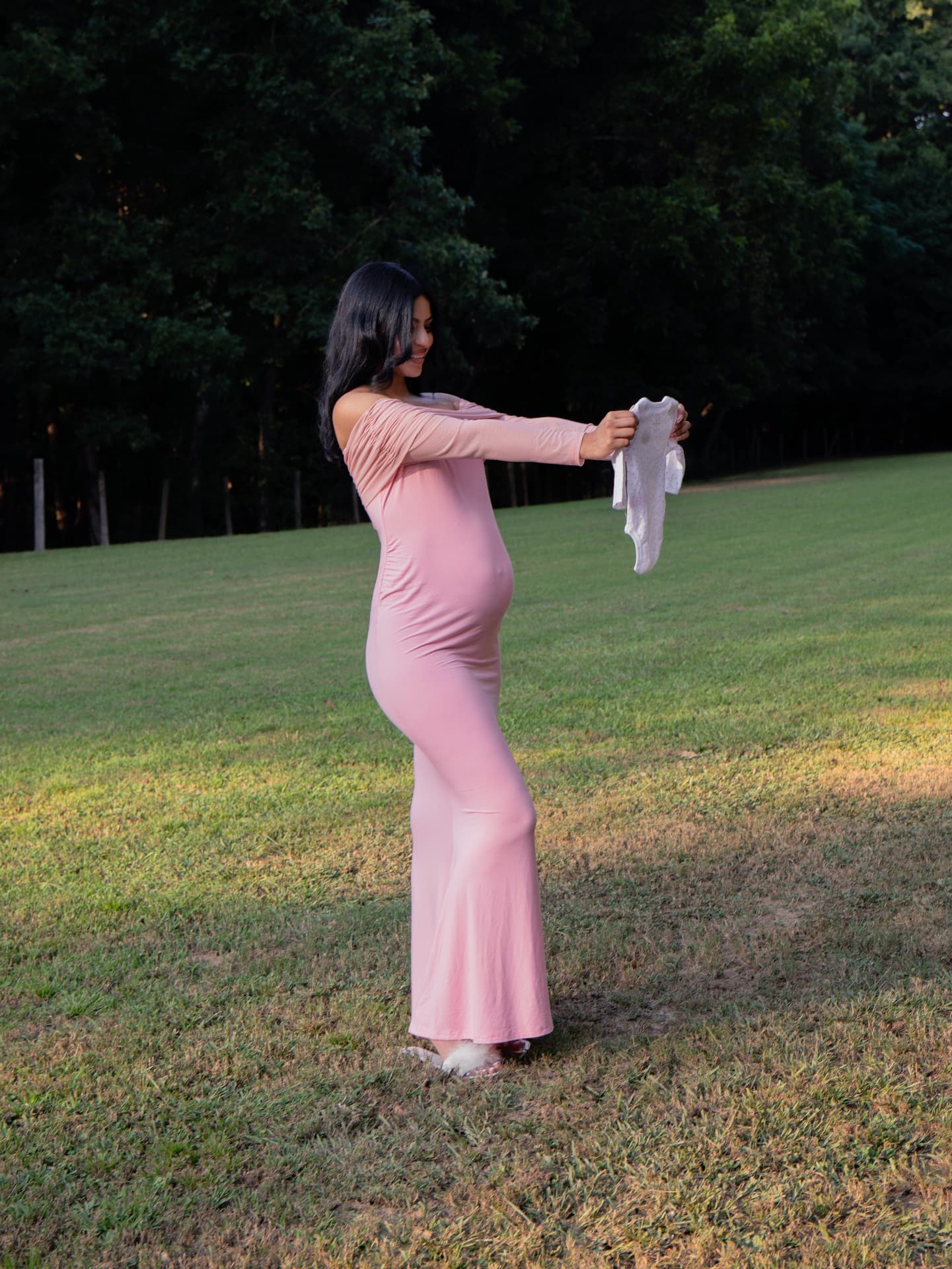 Expecting mother holding up a baby onesie in a sunlit meadow