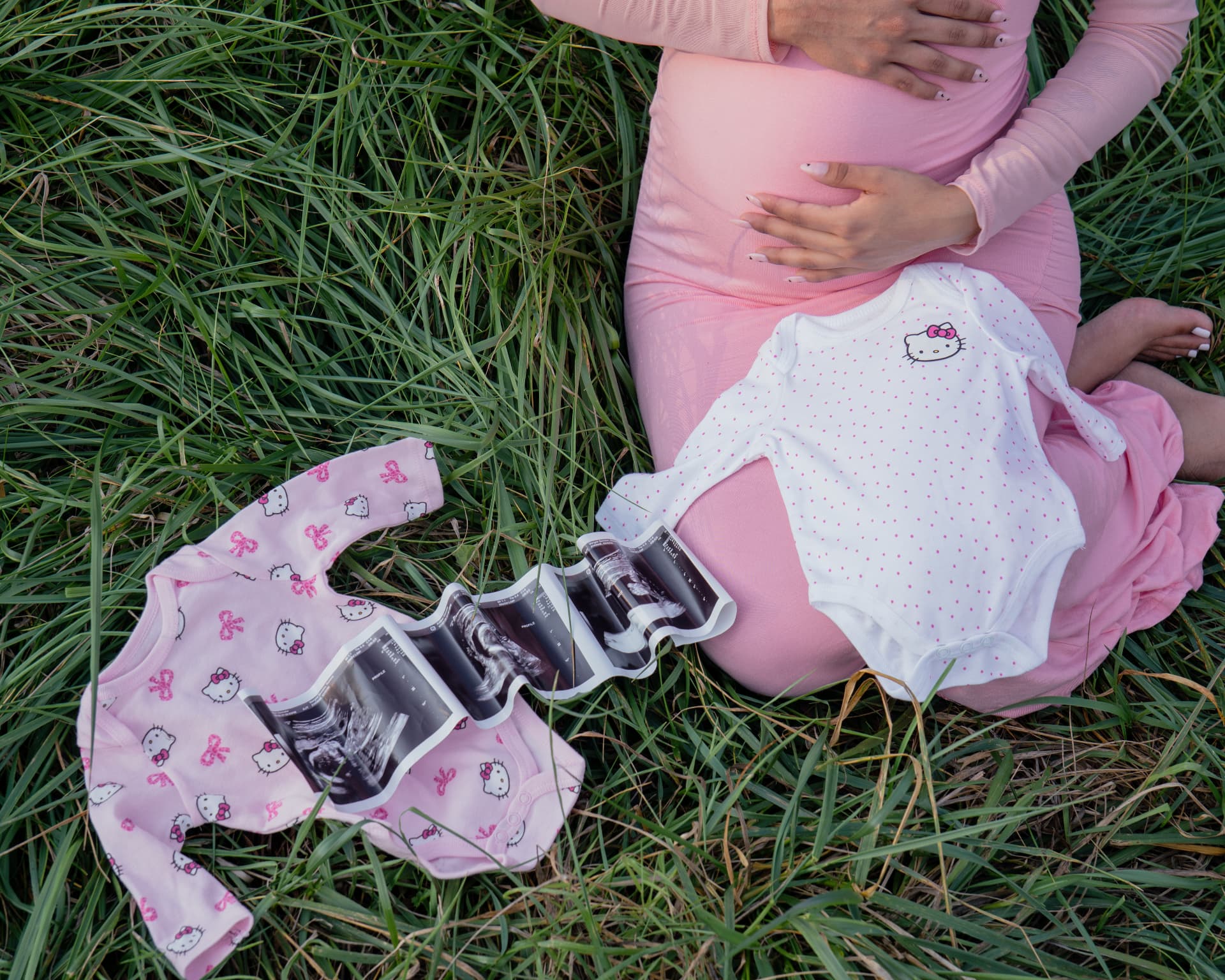Overhead maternity flat lay with baby onesies and ultrasound photos in the grass