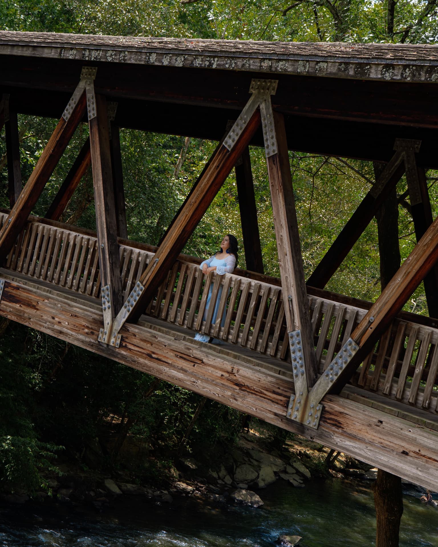Wide view of a woman in a blue gown standing on a covered bridge above a creek