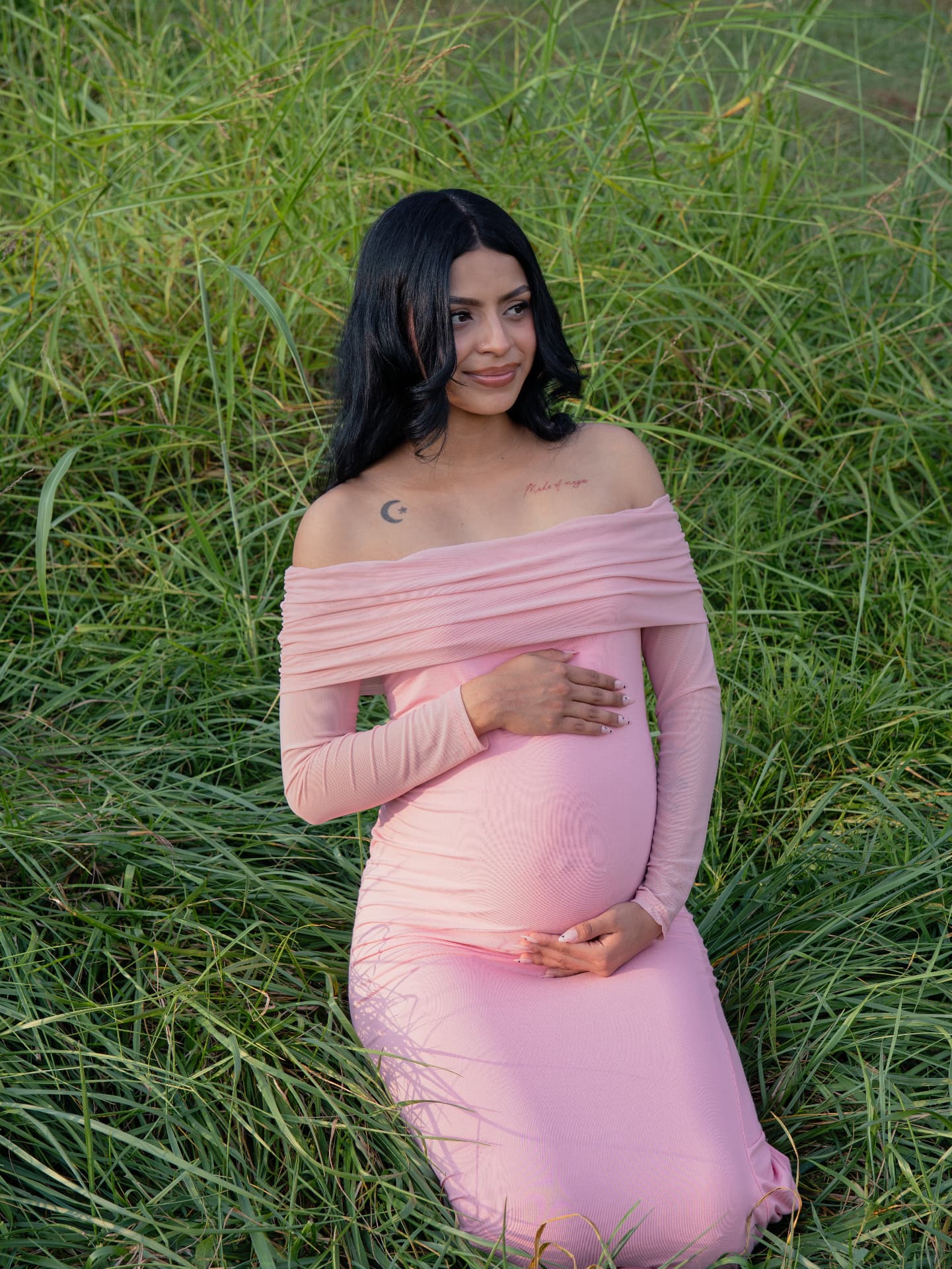 Mother-to-be in a pink gown seated in tall green grass at golden hour