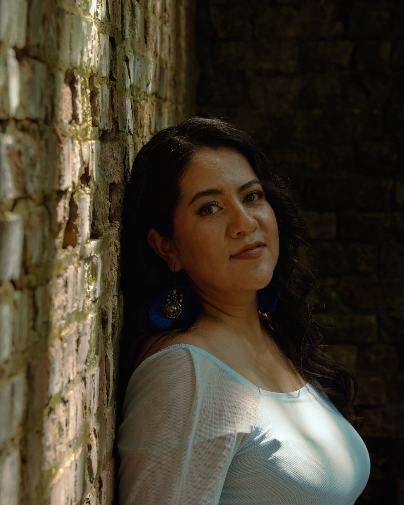 Close portrait of a woman with blue earrings leaning against a sunlit brick wall